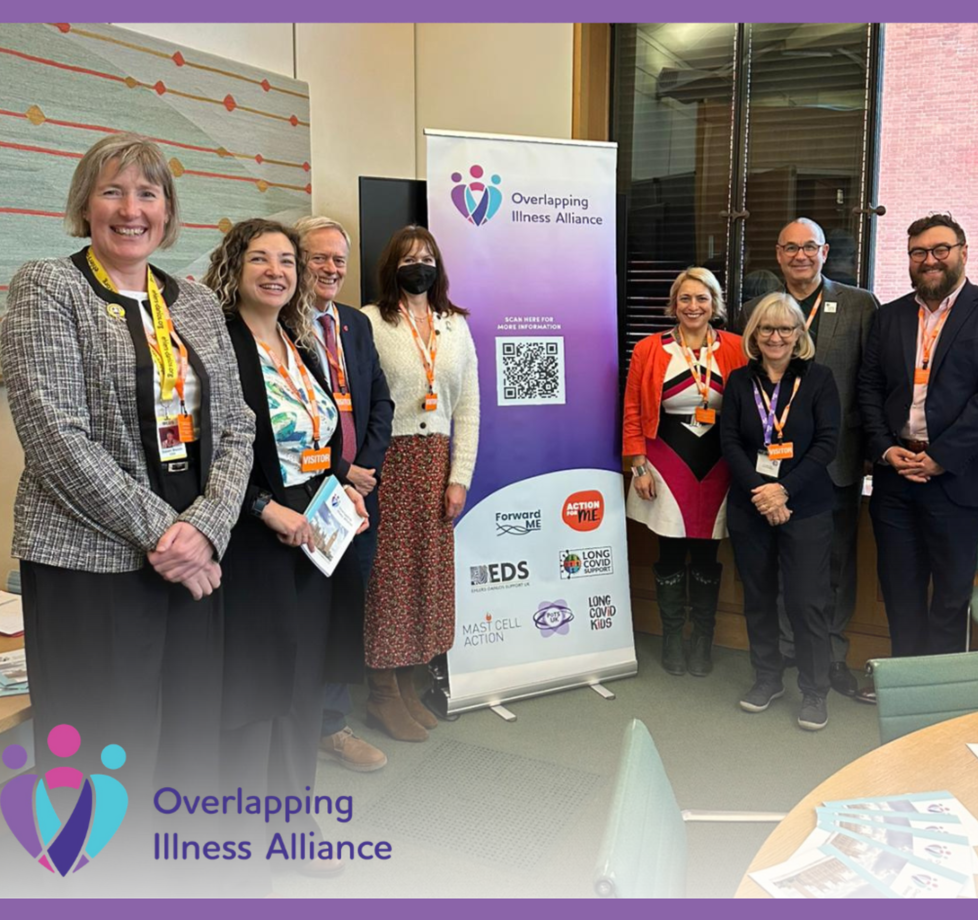 A group of nine adults stand smiling beside an Overlapping Illness Alliance banner in a parliamentary room. They wear visitor lanyards and hold leaflets. The banner shows the Alliance logo and partner organisations. A table with documents is visible in the foreground.