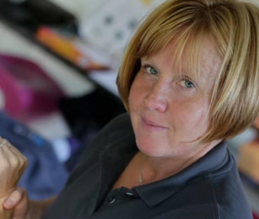 Photograph of Ellen Woodley, a woman with a blonde bob, wearing a dark blue t-shirt. She is looking directly at the camera, and the picture is taken at an angle, showing just her head and shoulders.