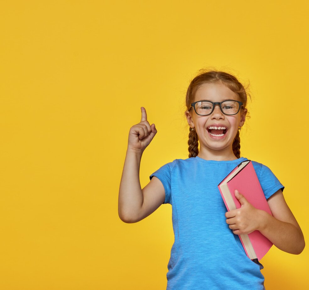A young girl wearing glasses, holding a book with her finger up in the air ready to ask a question